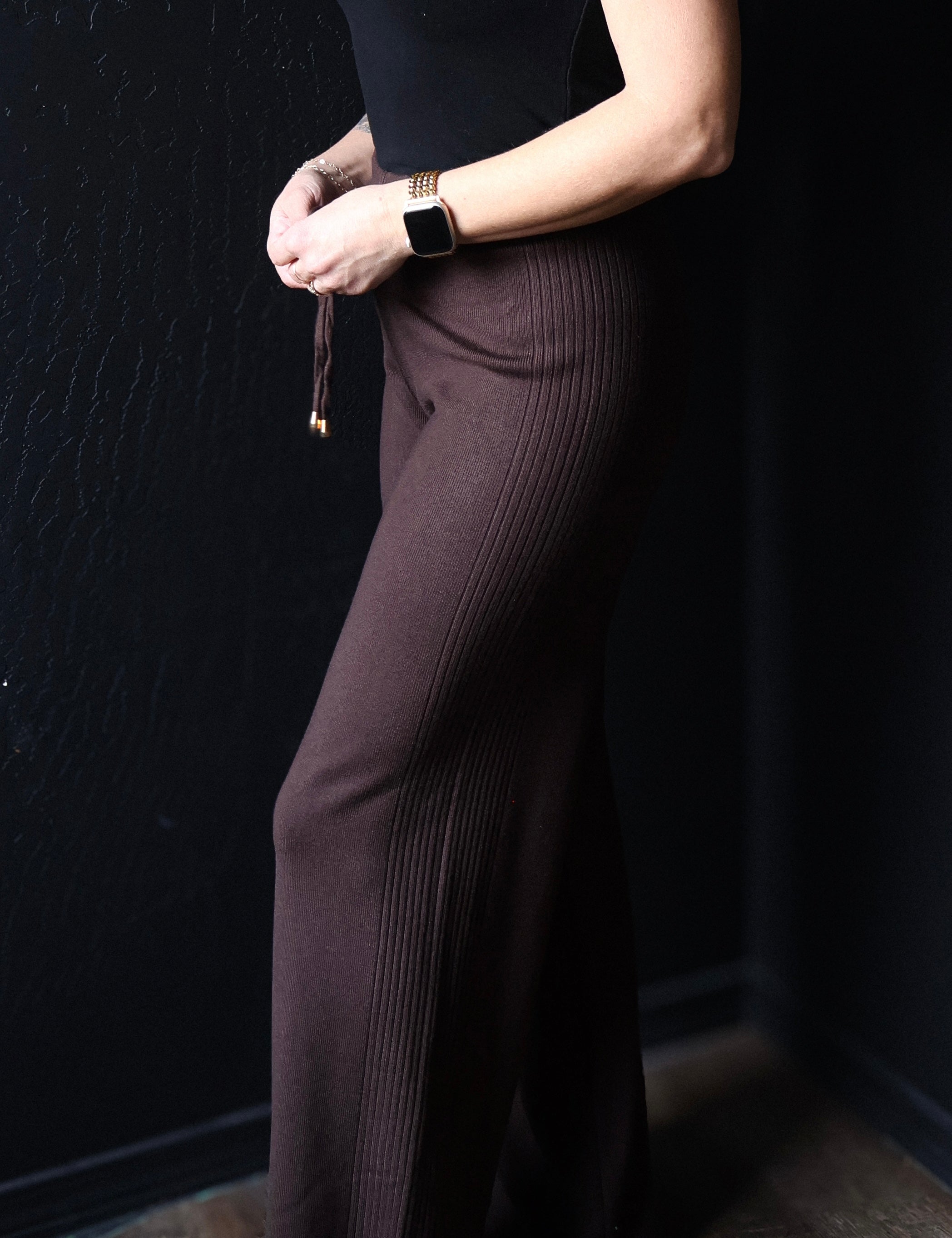 Person wearing a black top and brown ribbed pants against a dark background