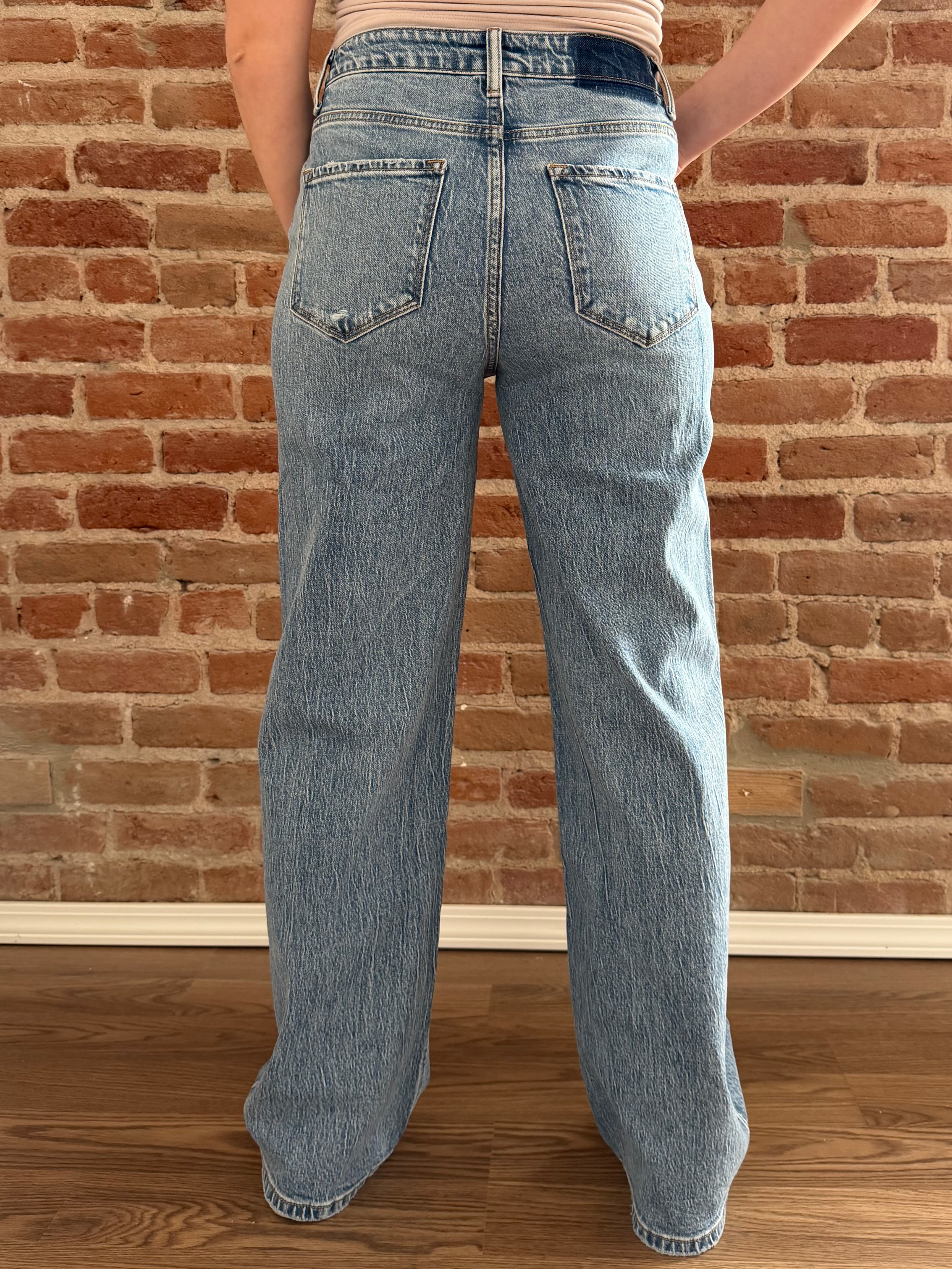 Person wearing light blue jeans standing against a brick wall.