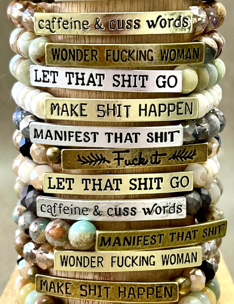 A stack of beaded bracelets with various etched metal bars, featuring different phrases such as 'MAKE SH*T HAPPEN' and 'WONDER F*CKING WOMAN'.