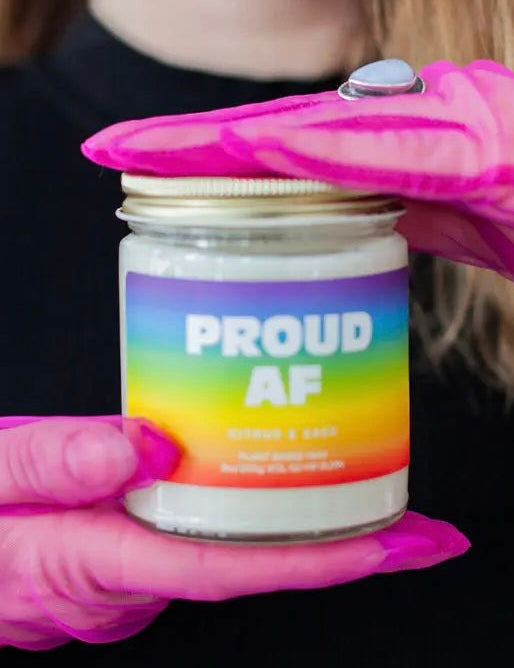 Jar labeled 'PROUD AF' with a colorful label held by pink gloves against a dark background