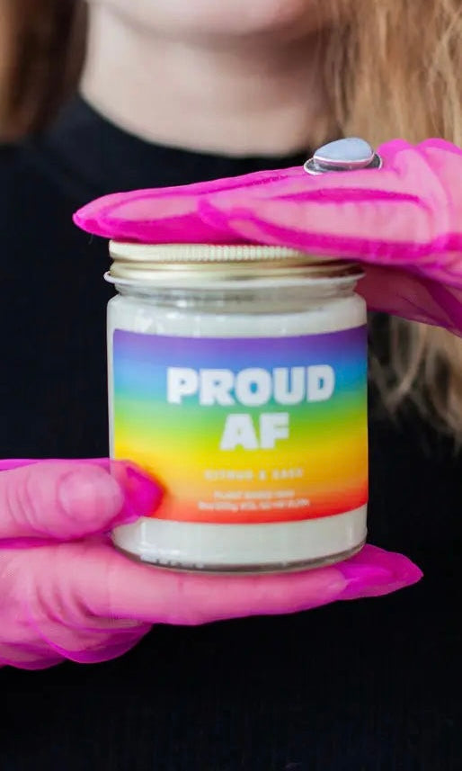 Jar labeled 'PROUD AF' with a colorful label held by pink gloves against a dark background