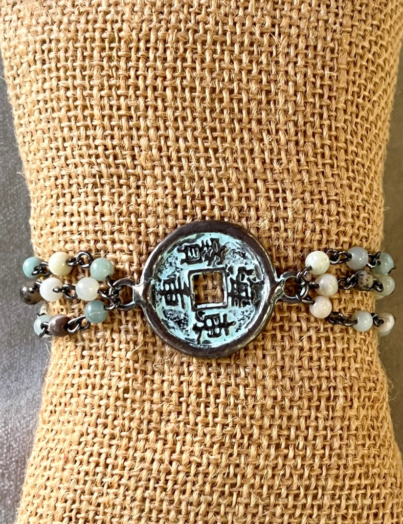 Amazonite beaded bracelet with a patina coin charm and black metal chain link, displayed on a burlap-textured surface.