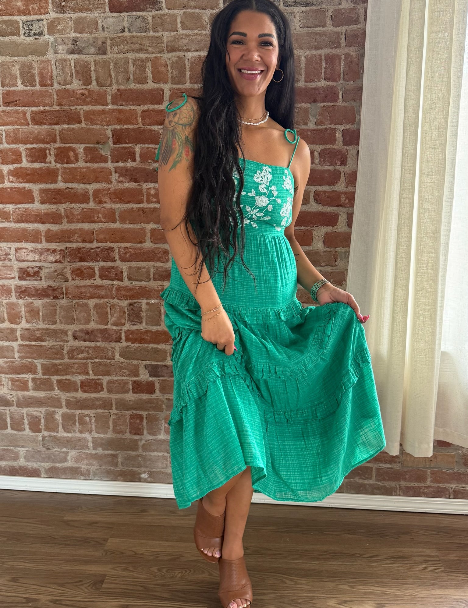Woman wearing a green dress with floral straps against a brick wall.