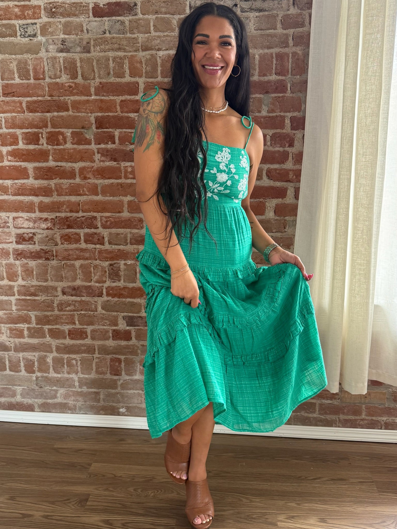 Woman wearing a green dress with floral straps against a brick wall.