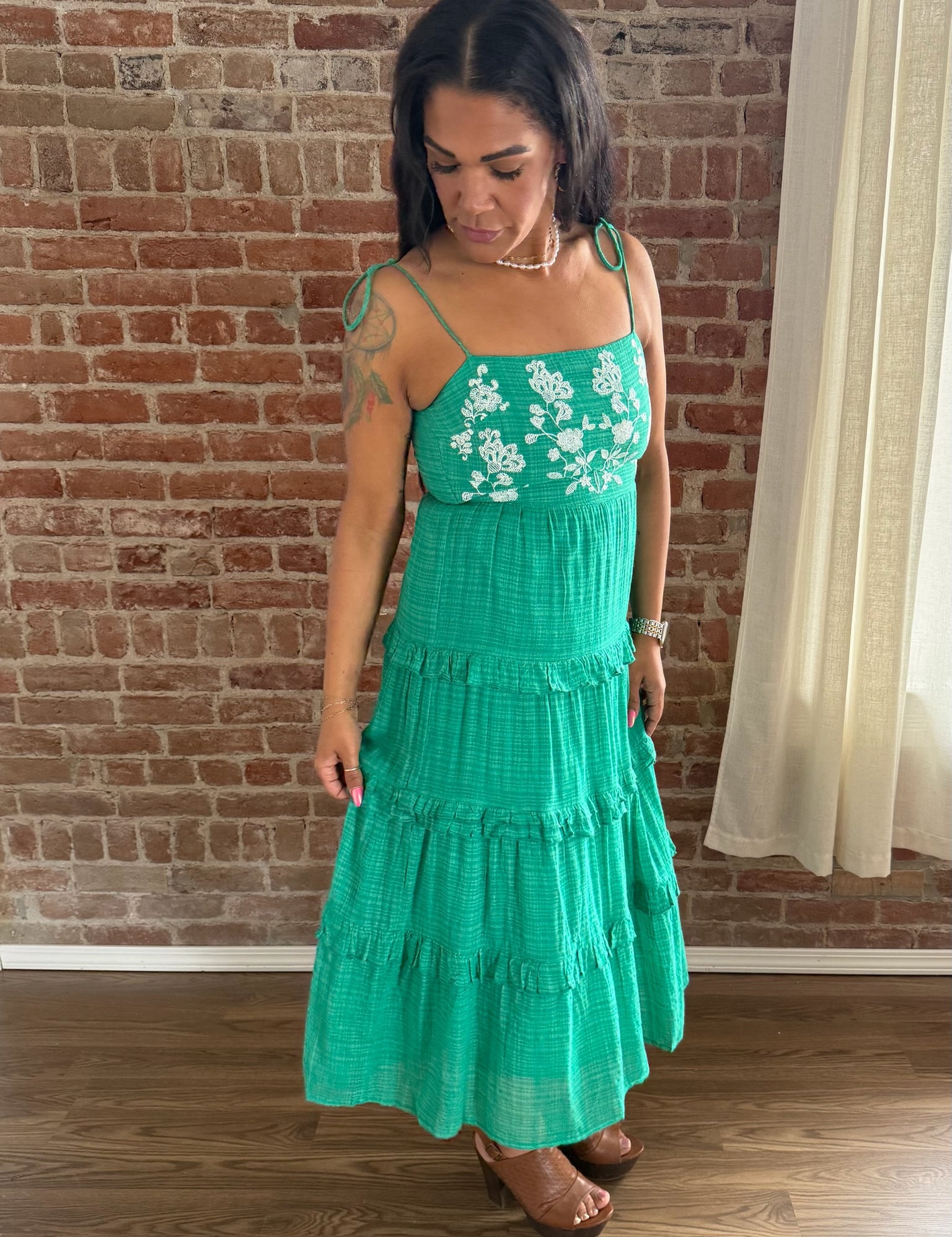 Woman wearing a green dress with floral embroidery in front of a brick wall.