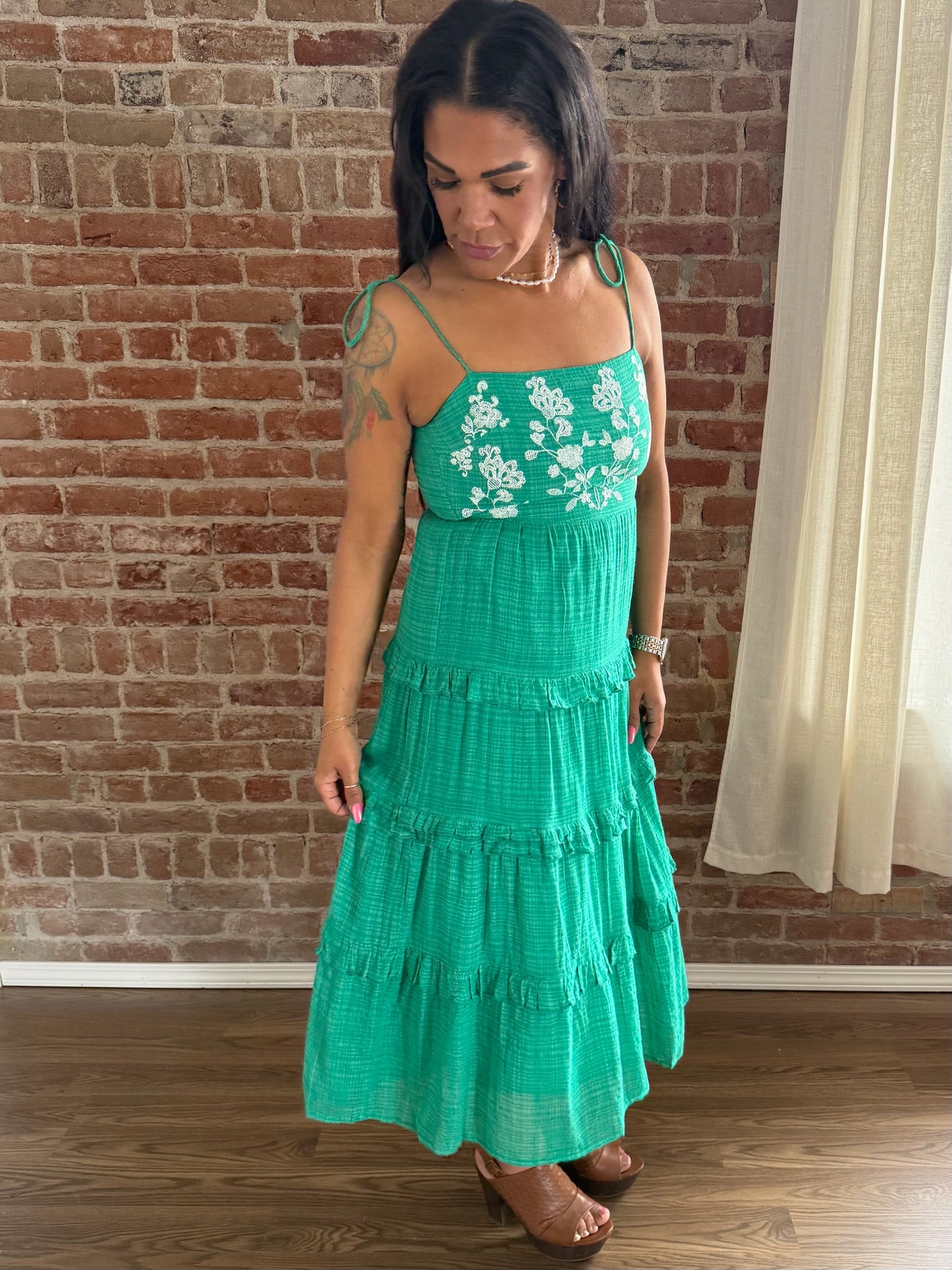 Woman wearing a green dress with floral embroidery in front of a brick wall.
