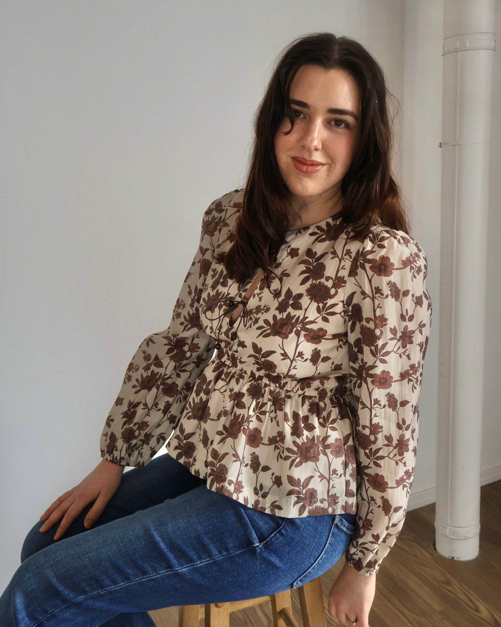 Woman wearing a floral blouse and jeans sitting on a stool indoors.