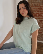 Woman wearing a light green patterned shirt sitting against a brick wall.