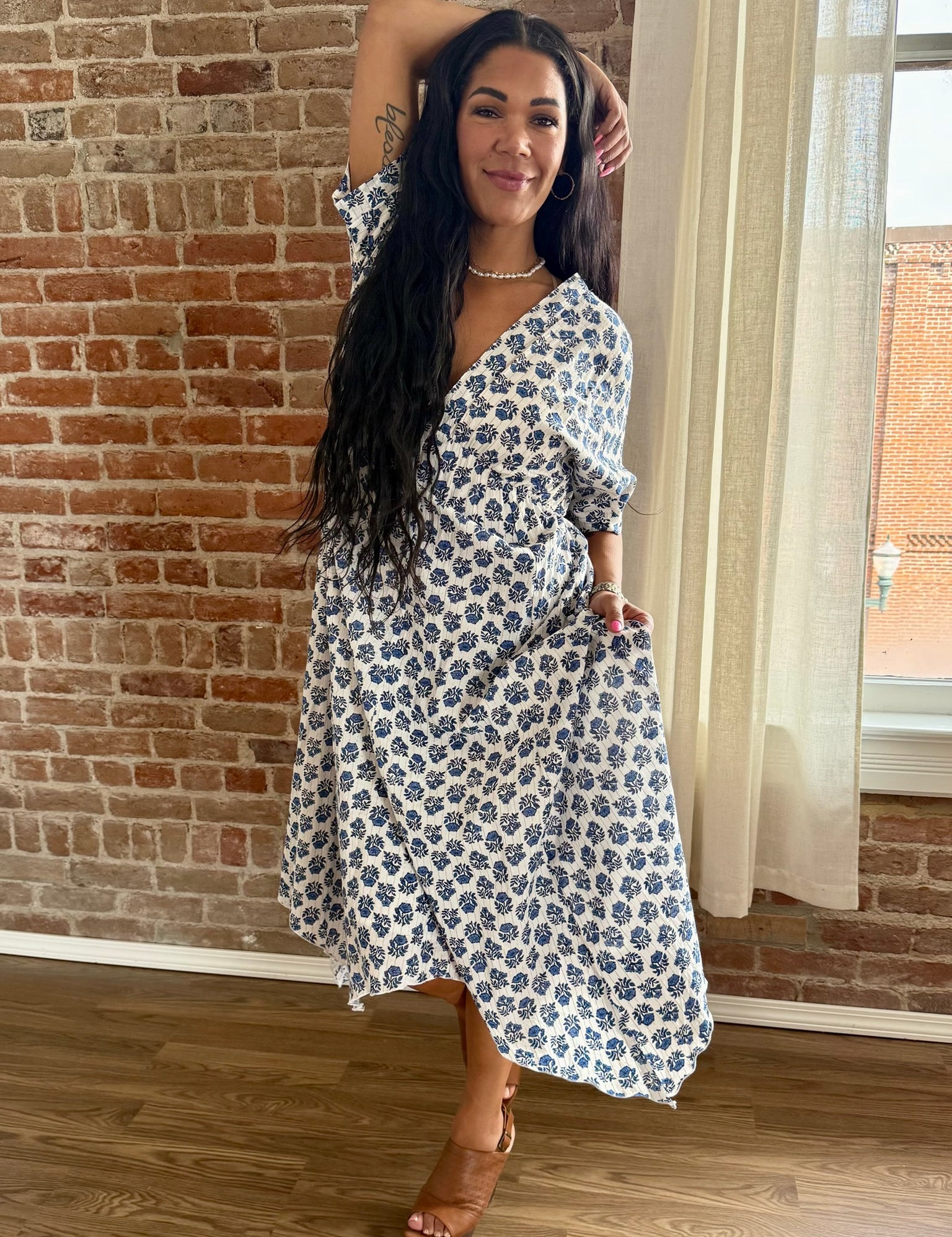 Woman in a floral dress standing against a brick wall indoors.