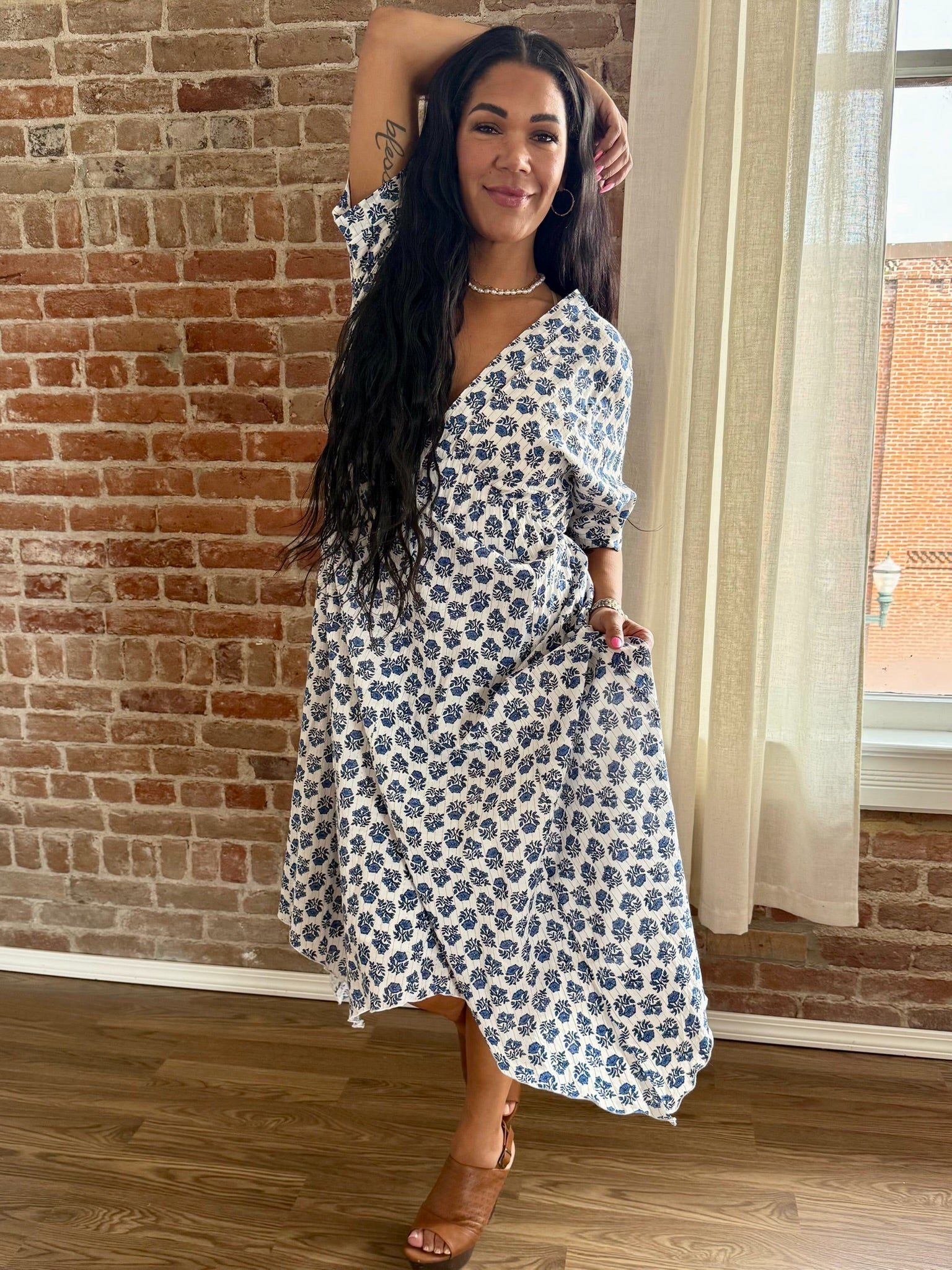 Woman in a floral dress standing against a brick wall indoors.